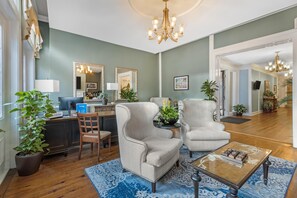 Ein Wohnzimmer mit einer Couch, Sesseln, einem Couchtisch und einer Chandelier. Ein Schreibtisch mit Computer und Pflanze ist ebenfalls vorhanden.