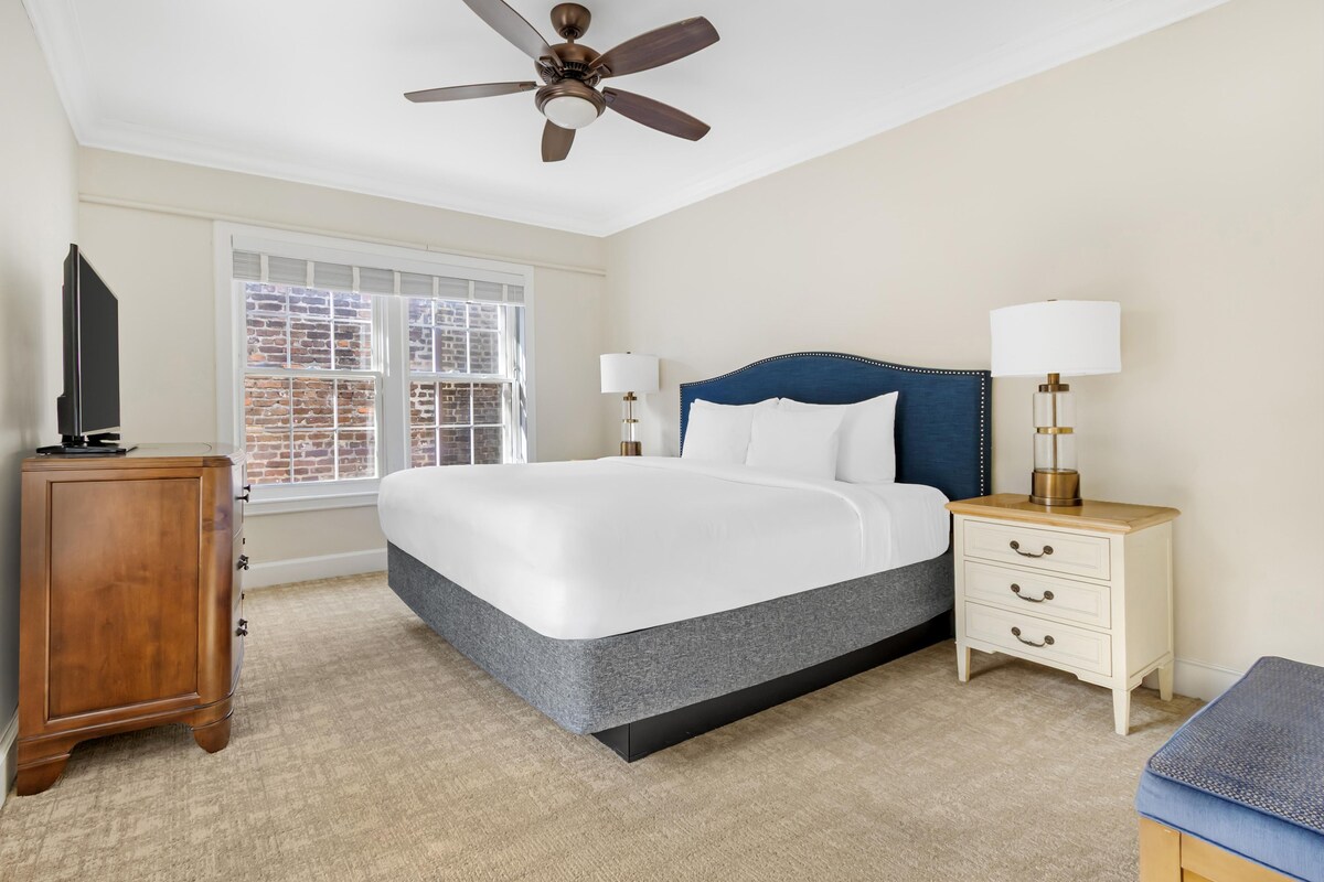 A bedroom with a bed, nightstands, a ceiling fan, a window with a view of a brick building, and a wooden cabinet.