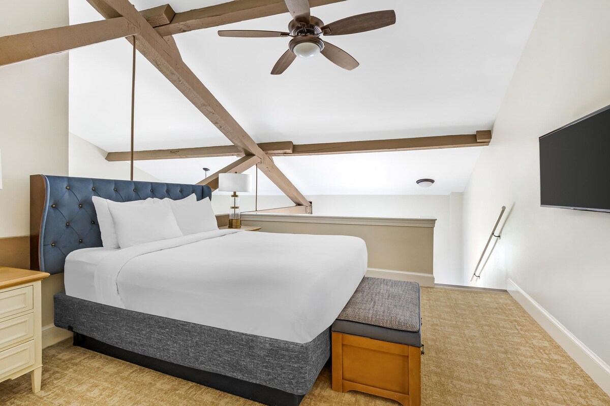 A bedroom with a bed, wooden beams, a ceiling fan, a nightstand, a bedside table, a lamp, and a TV mounted on the wall.