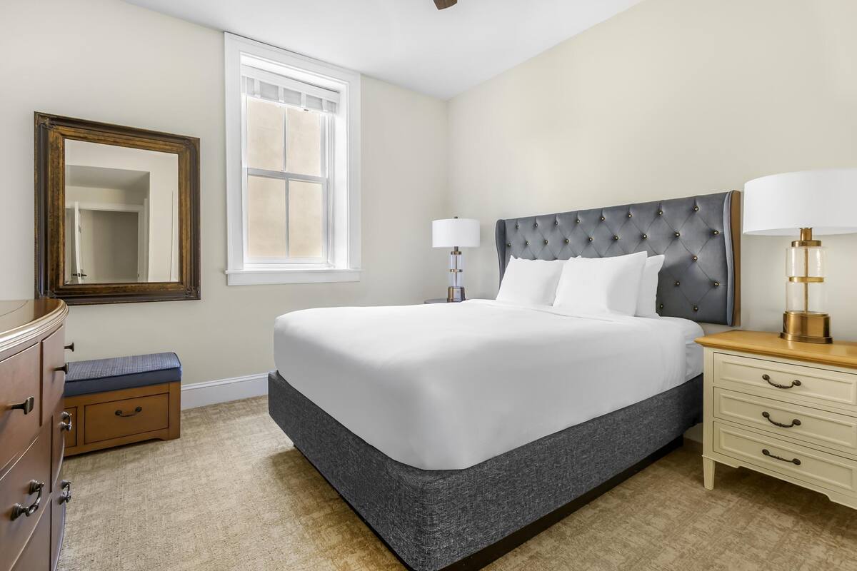 A neatly arranged bedroom with a large bed, a bedside table with a lamp, a dresser, and a mirror.