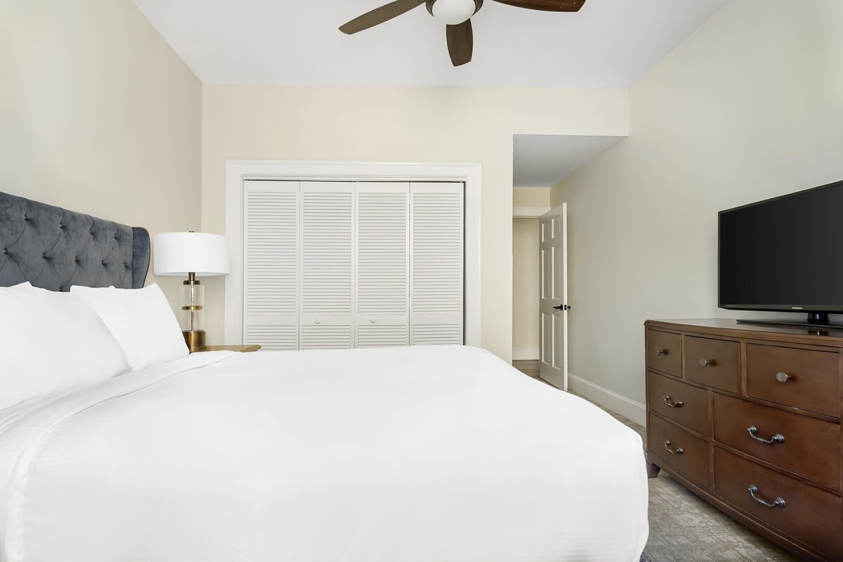 A bedroom with a bed, a television, a dresser, a ceiling fan, and a closet.