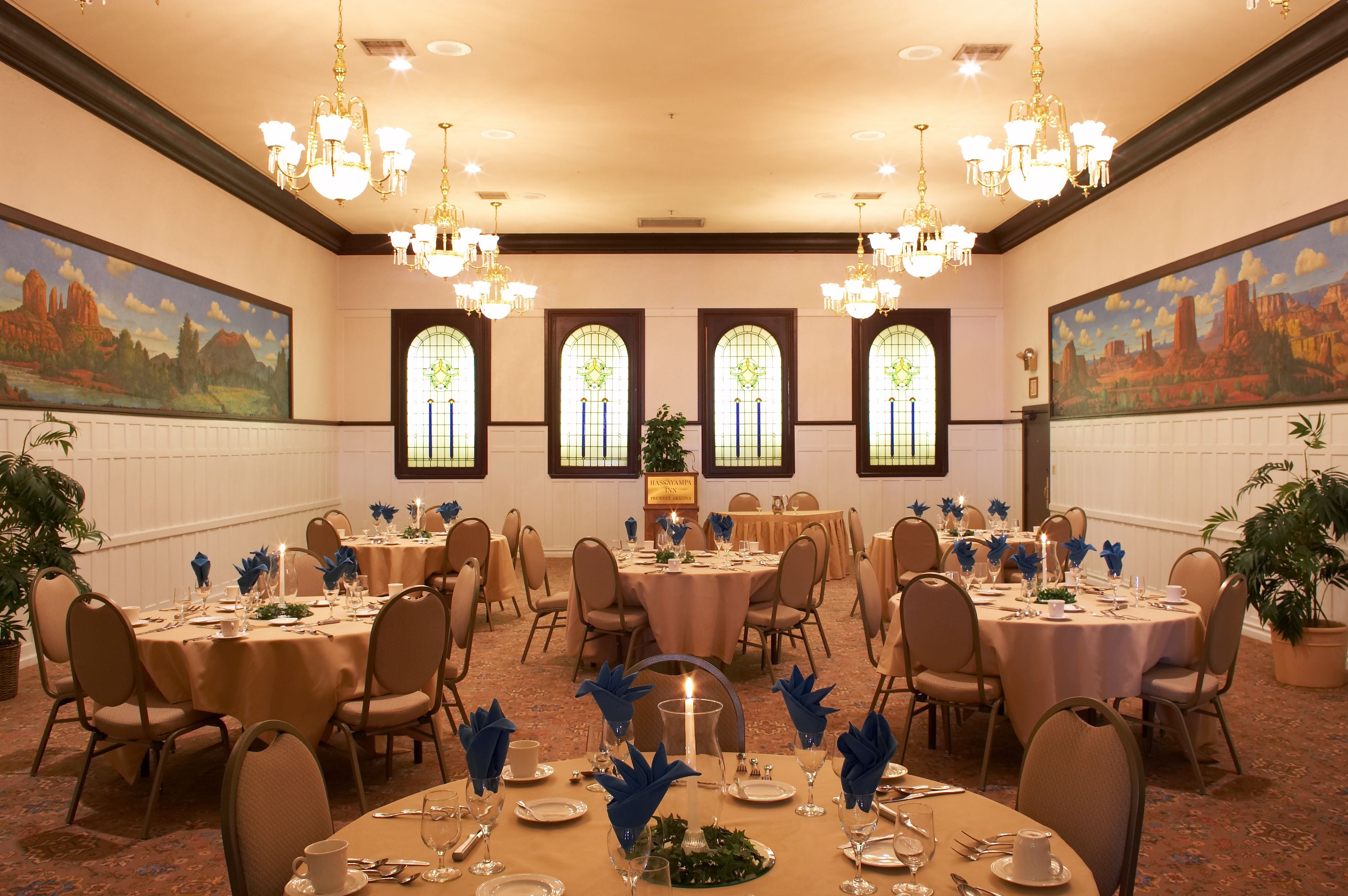 Un salón de banquetes con mesas redondas, sillas, candelabros y grandes ventanales con vidrieras de colores.