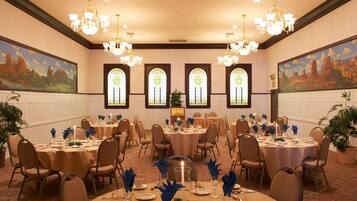 A banquet hall with round tables, chairs, chandeliers, and large windows with stained glass.