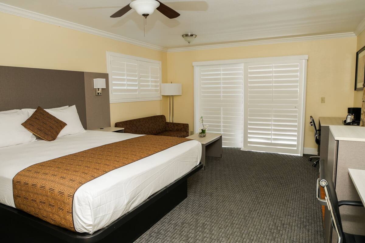 A hotel room with a large bed, a desk, a chair, a ceiling fan, and a window with blinds.