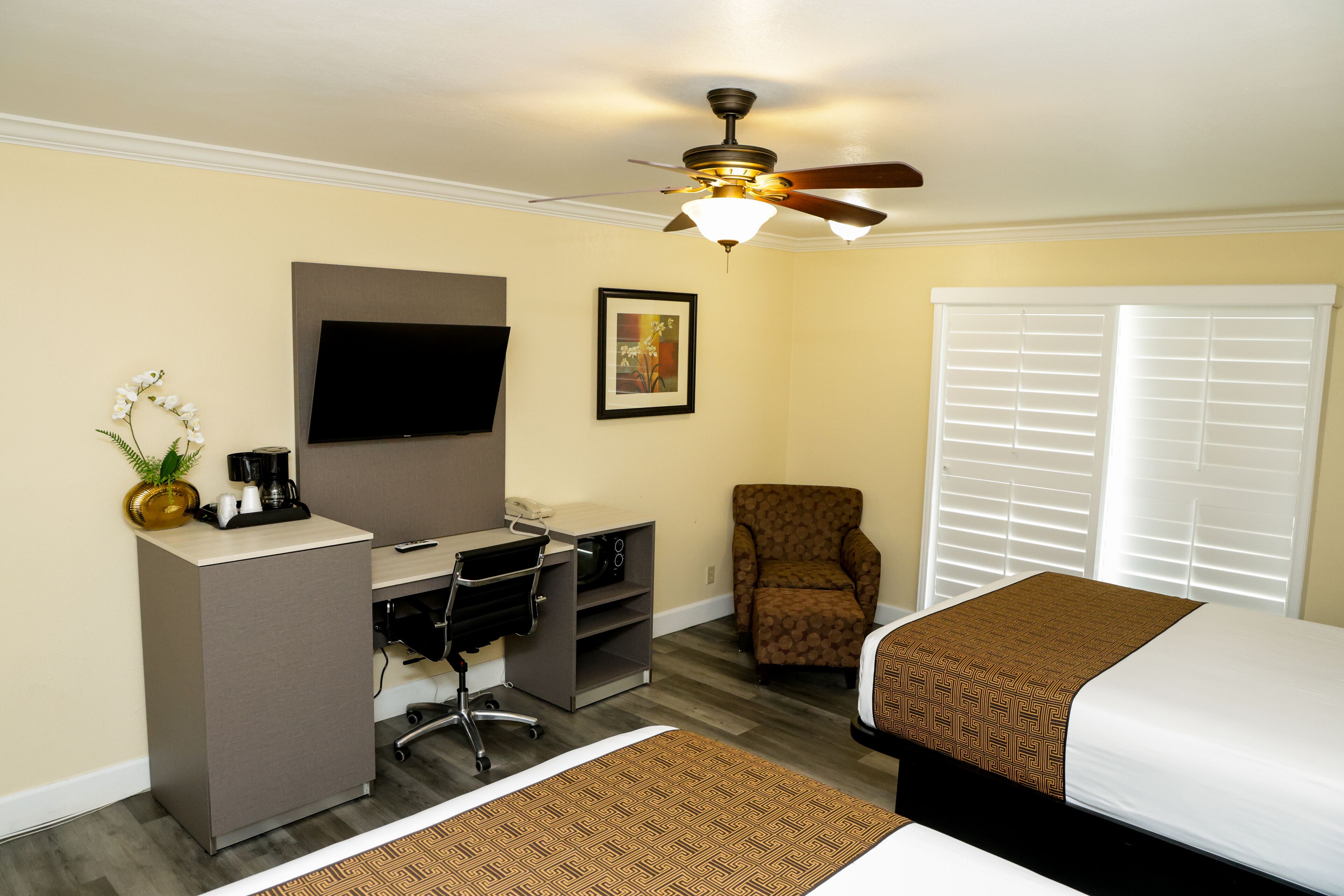 A hotel room with a bed, desk, chair, television, ceiling fan, and window with blinds.