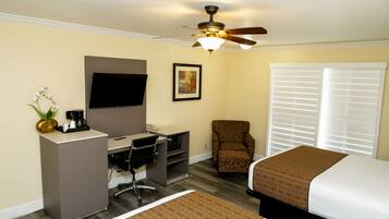 A hotel room with a bed, desk, chair, television, ceiling fan, and window with blinds.