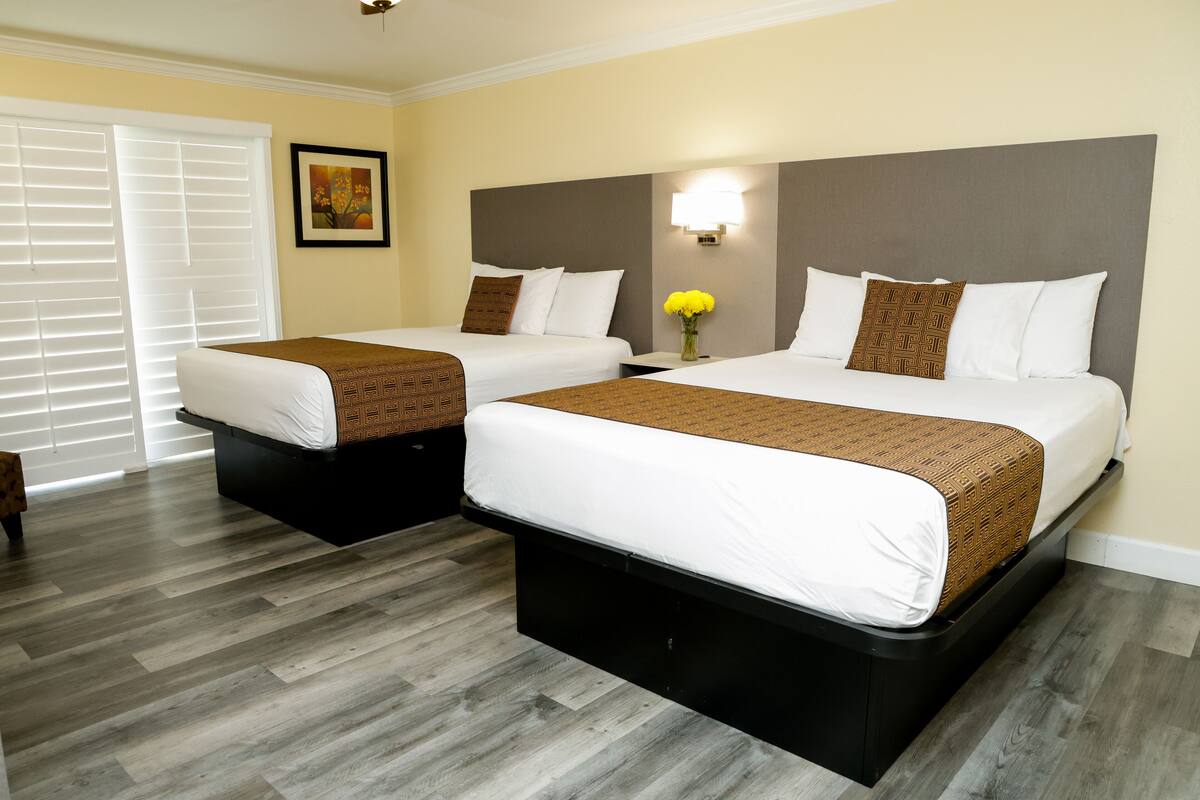 A hotel room with two beds, a large window with blinds, a wall-mounted light, and a framed picture.