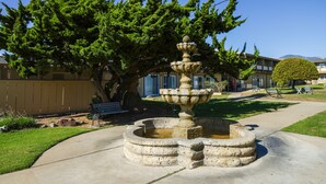 A stone fountain surrounded by a circular stone border, a tree, and a bench in a park-like setting. - Silver Surf Motel (San Simeon)