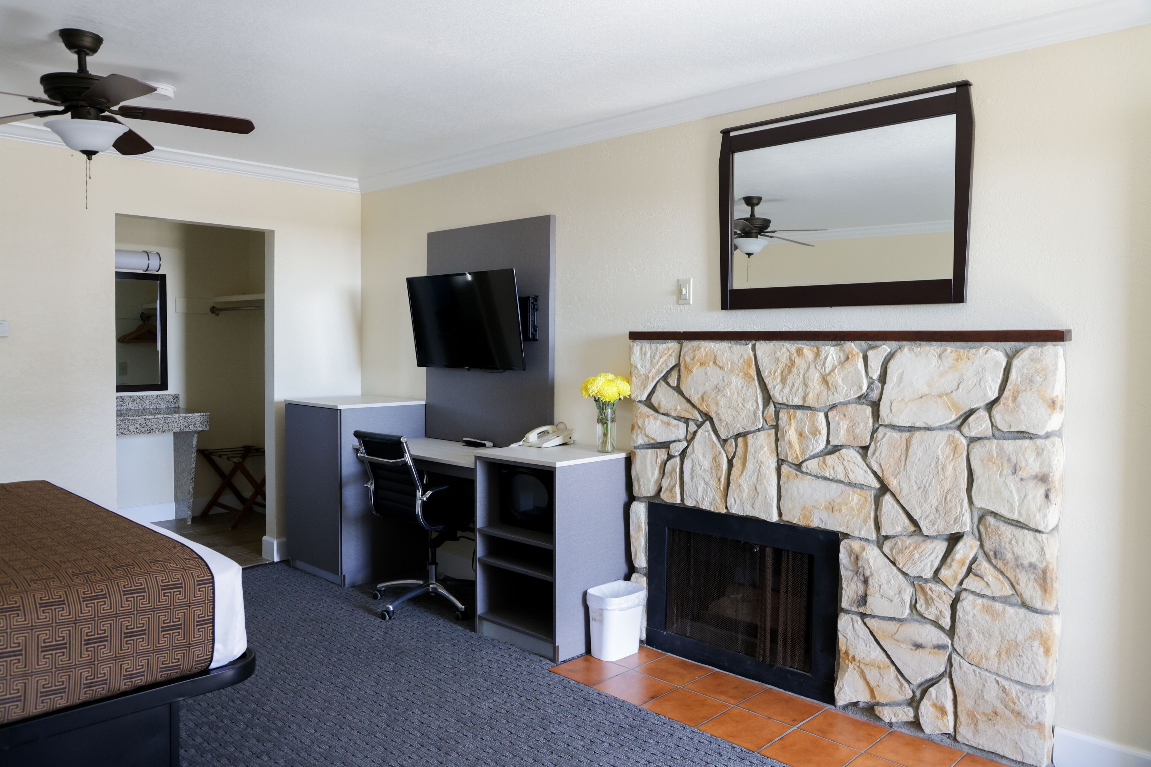 A hotel room with a bed, desk, chair, TV, fireplace, and a ceiling fan.