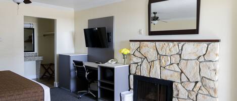 A hotel room with a bed, desk, chair, TV, fireplace, and a ceiling fan.