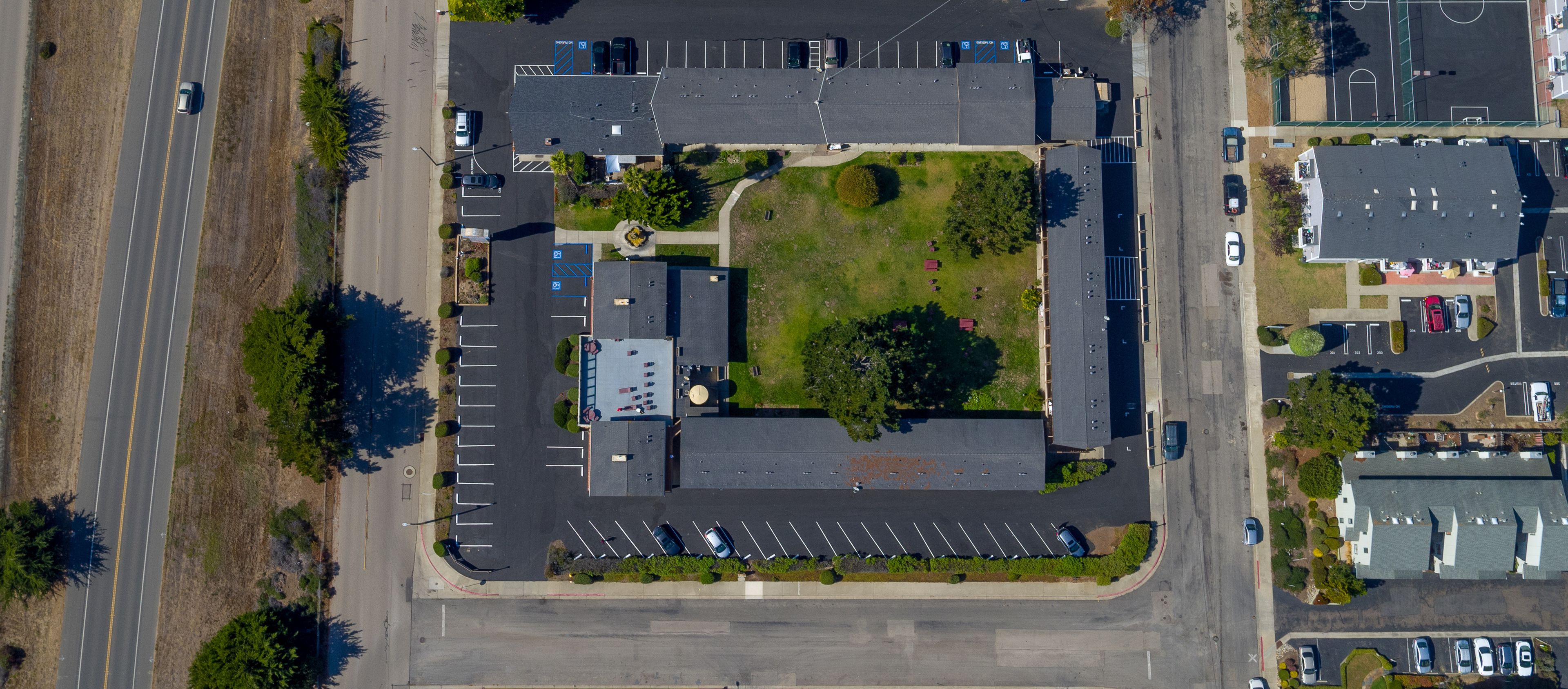 An aerial view of a parking lot with multiple rows of parked cars, a central grassy area, and surrounding buildings.