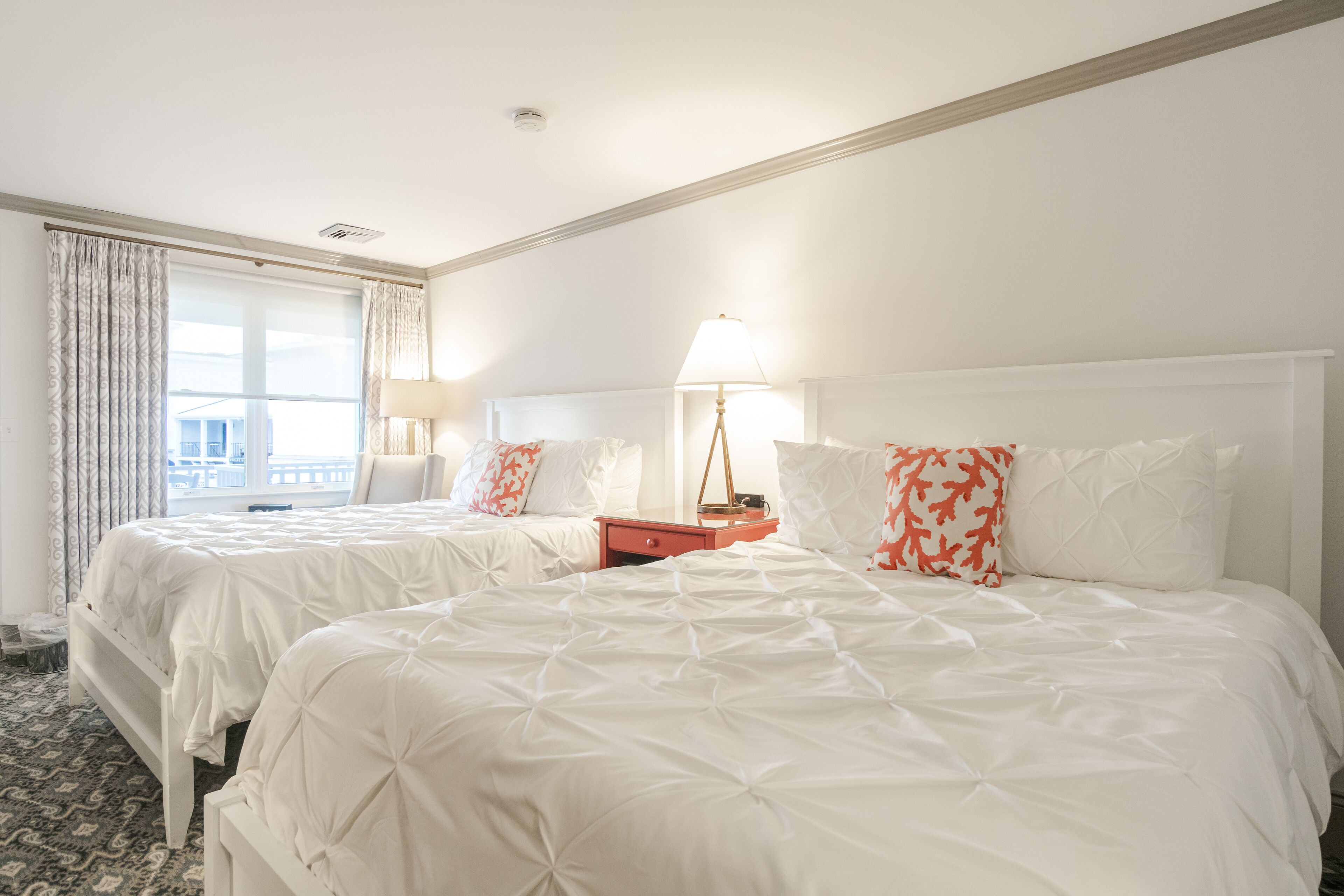 A hotel room with two beds, a nightstand, a lamp, and a window with curtains.