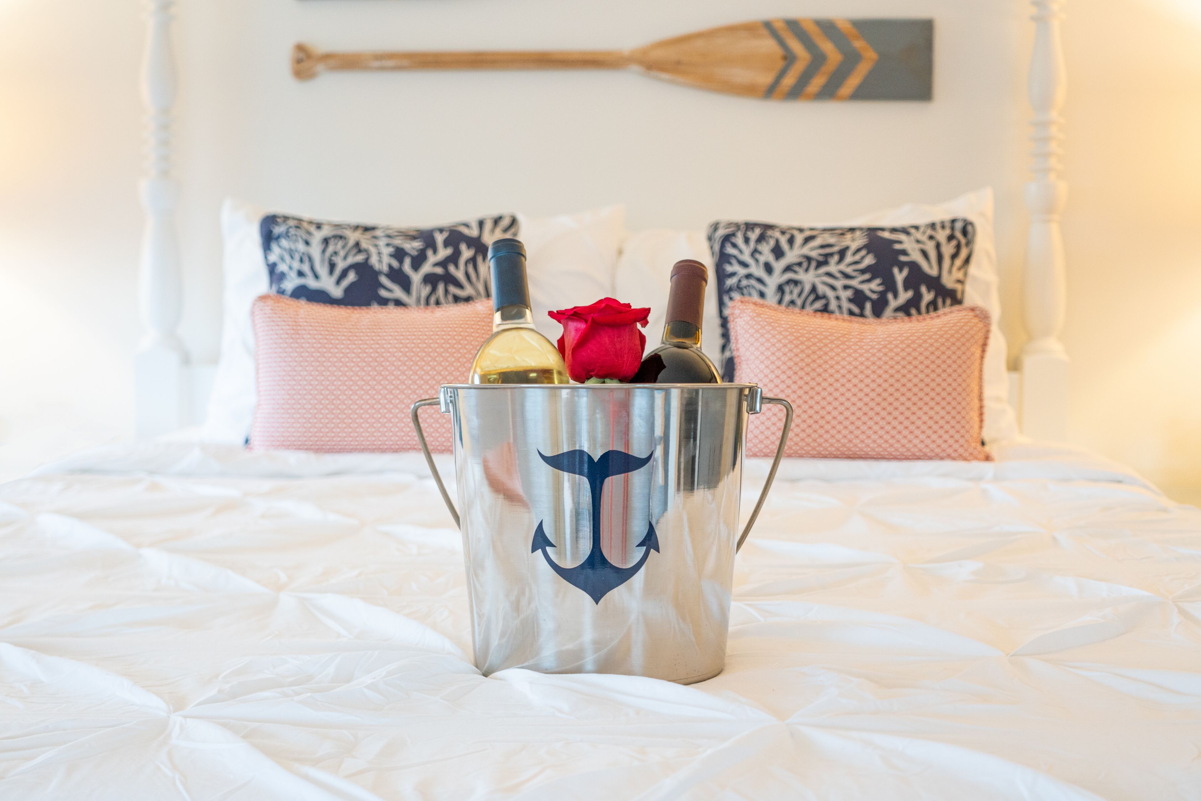 A bed with a white comforter, a metal bucket with a nautical anchor design, and two bottles with a red rose.