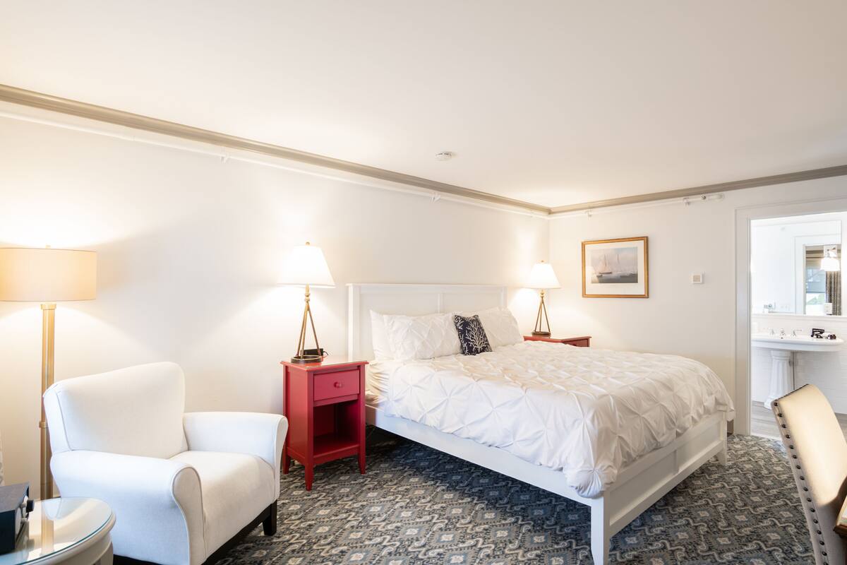 Une chambre d’hôtel avec un grand lit, une table de chevet rouge, un fauteuil blanc et une salle de bain visible à travers une porte ouverte.