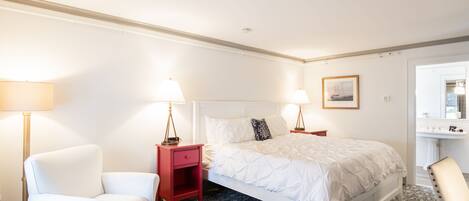 A hotel room with a large bed, a red nightstand, a white armchair, and a bathroom visible through an open door.