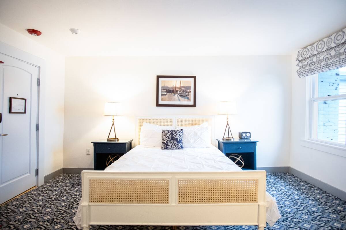 Une chambre à coucher bien rangée, avec un lit, deux tables de chevet, un tableau encadré au mur et une fenêtre avec des rideaux.