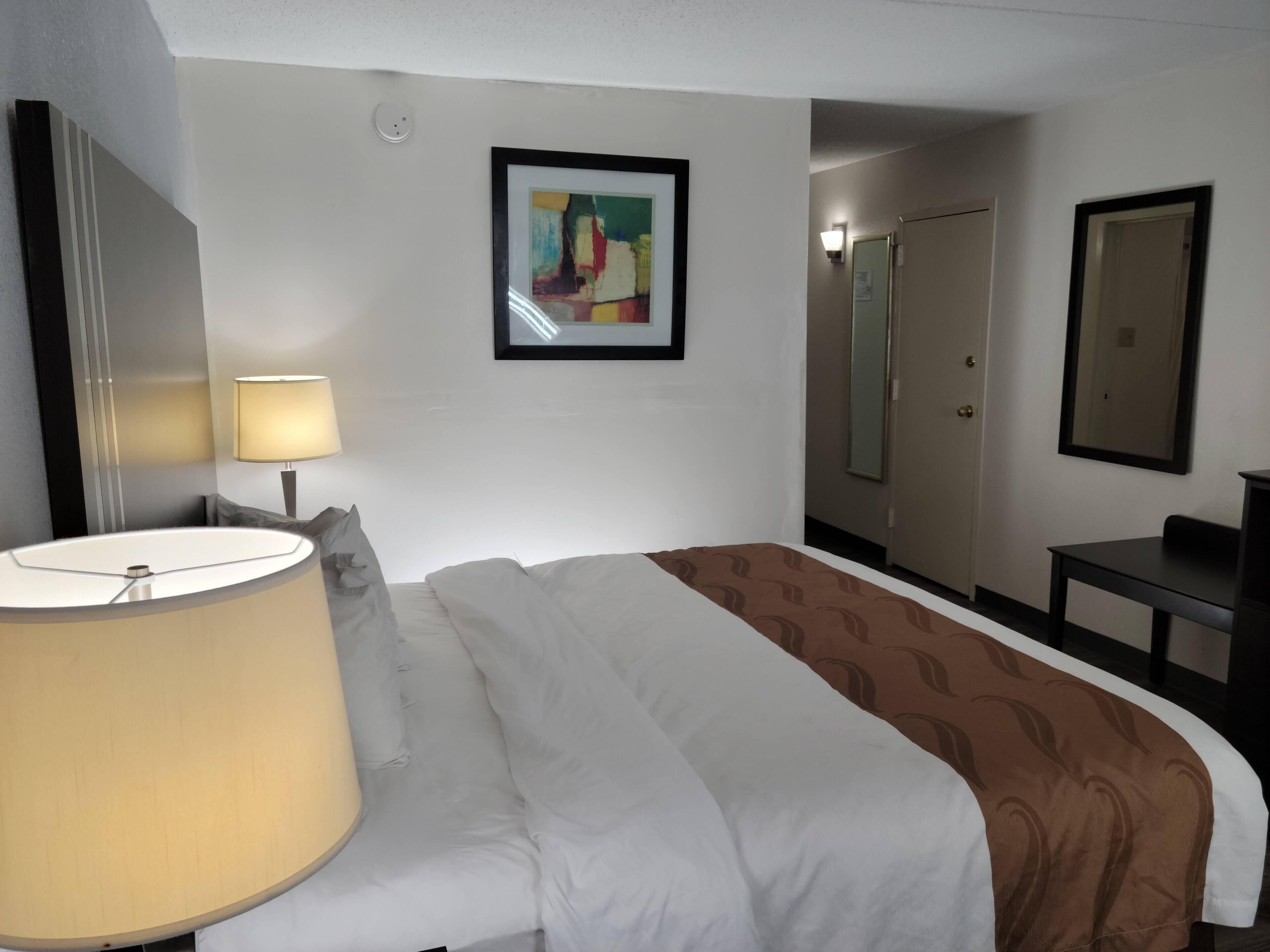 A hotel room with a bed, a bedside lamp, a framed abstract painting, a mirror, and a wardrobe.