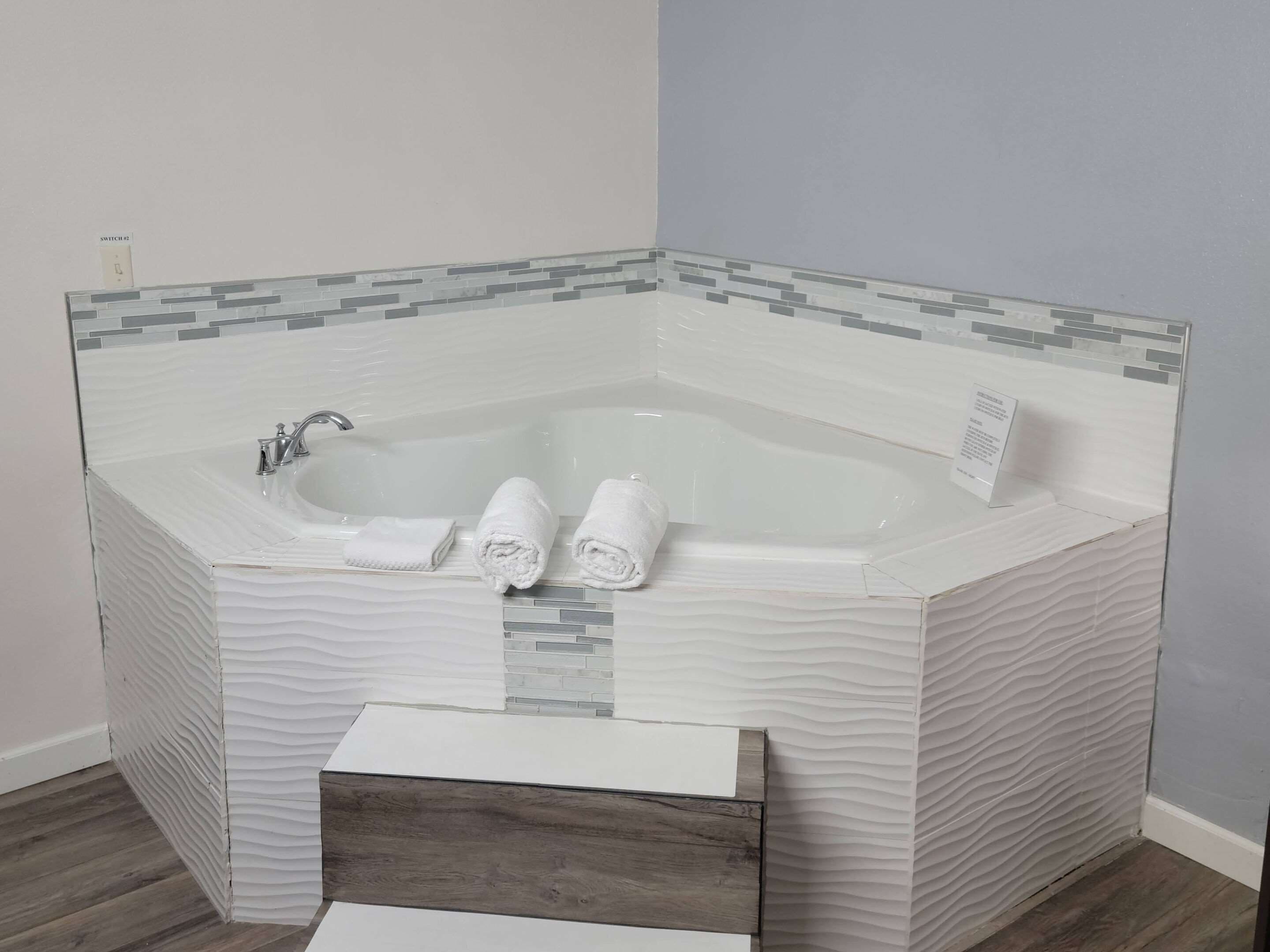 Ein Badezimmer im Eck mit einer in Glas eingefassten Badewanne, zwei aufgerollten Handtüchern und einer hölzernen Sitzbank.