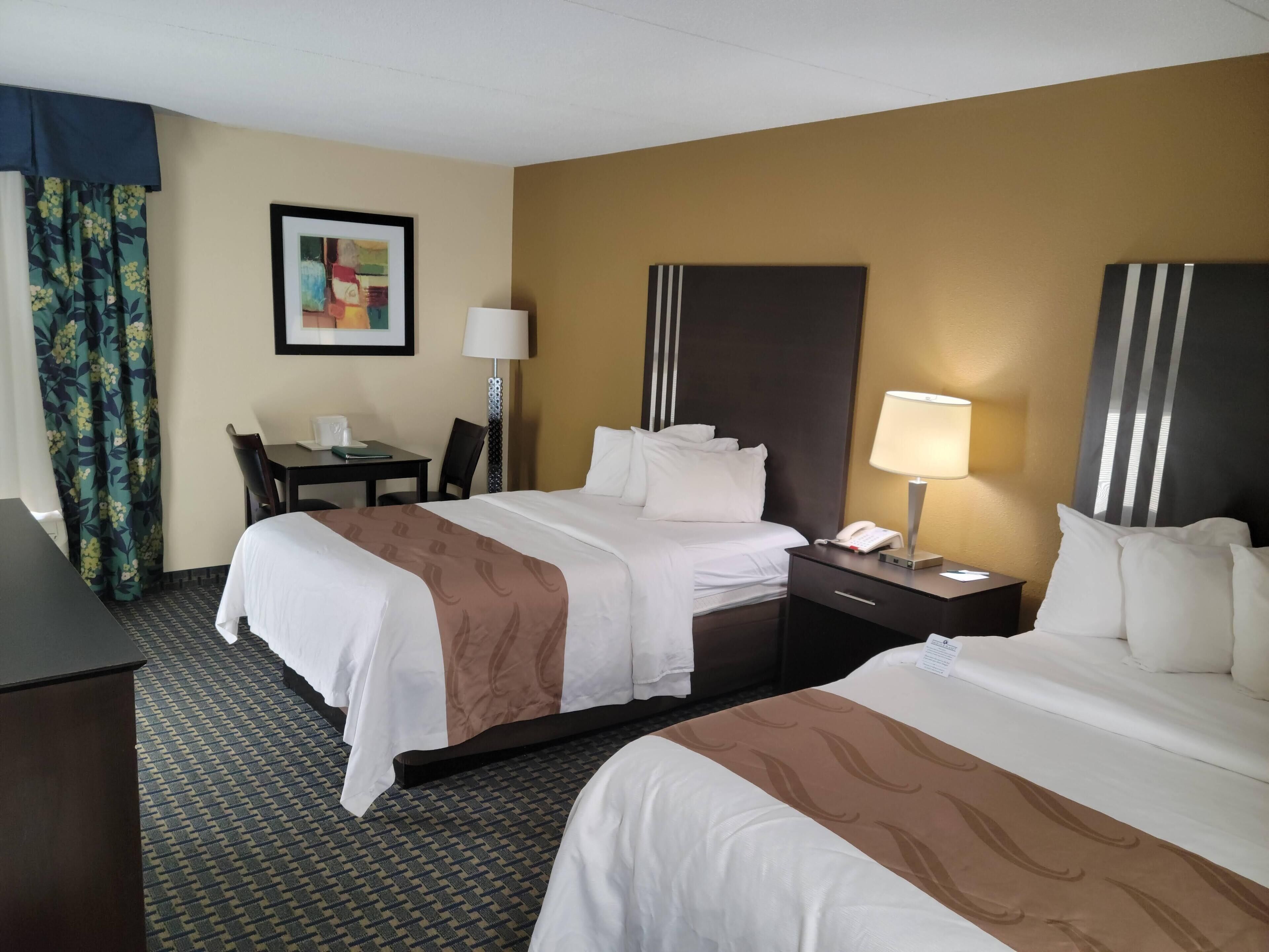A hotel room with two beds, a desk, a chair, a lamp, and a framed picture on the wall.