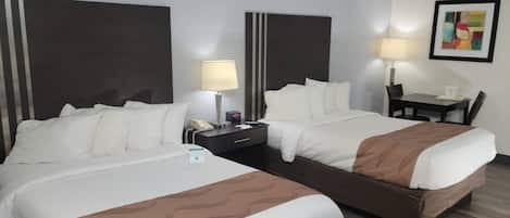 Ein Hotelzimmer mit zwei Betten, einem Schreibtisch, einem Stuhl, einer Lampe und einem gerahmten Bild an der Wand.