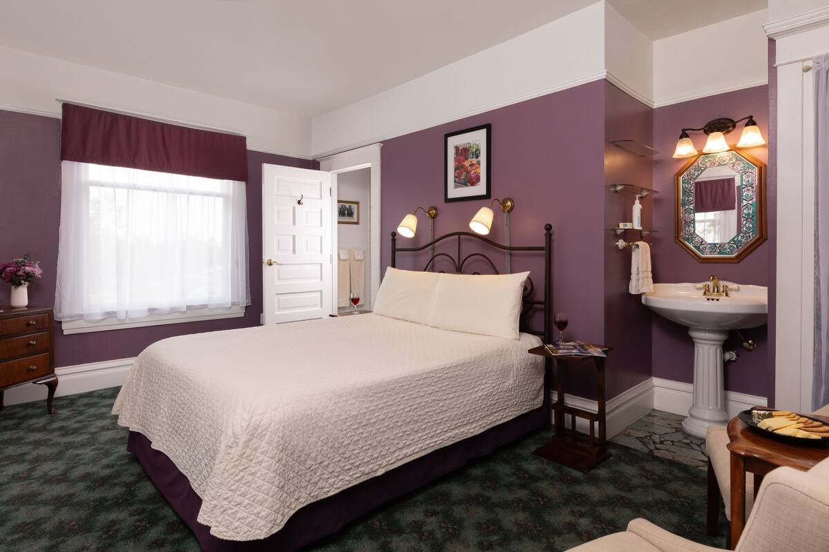 Une chambre à coucher comprenant un lit, une table de chevet, une commode, une fenêtre avec des rideaux, une salle de bain avec un lavabo et un miroir, et un tableau encadré accroché au mur.