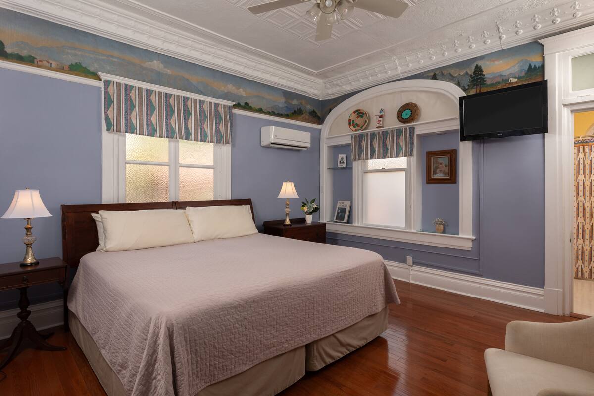 Une chambre à coucher comprenant un lit, deux tables de chevet avec des lampes, une fenêtre avec des rideaux, un téléviseur fixé au mur et un miroir décoratif.