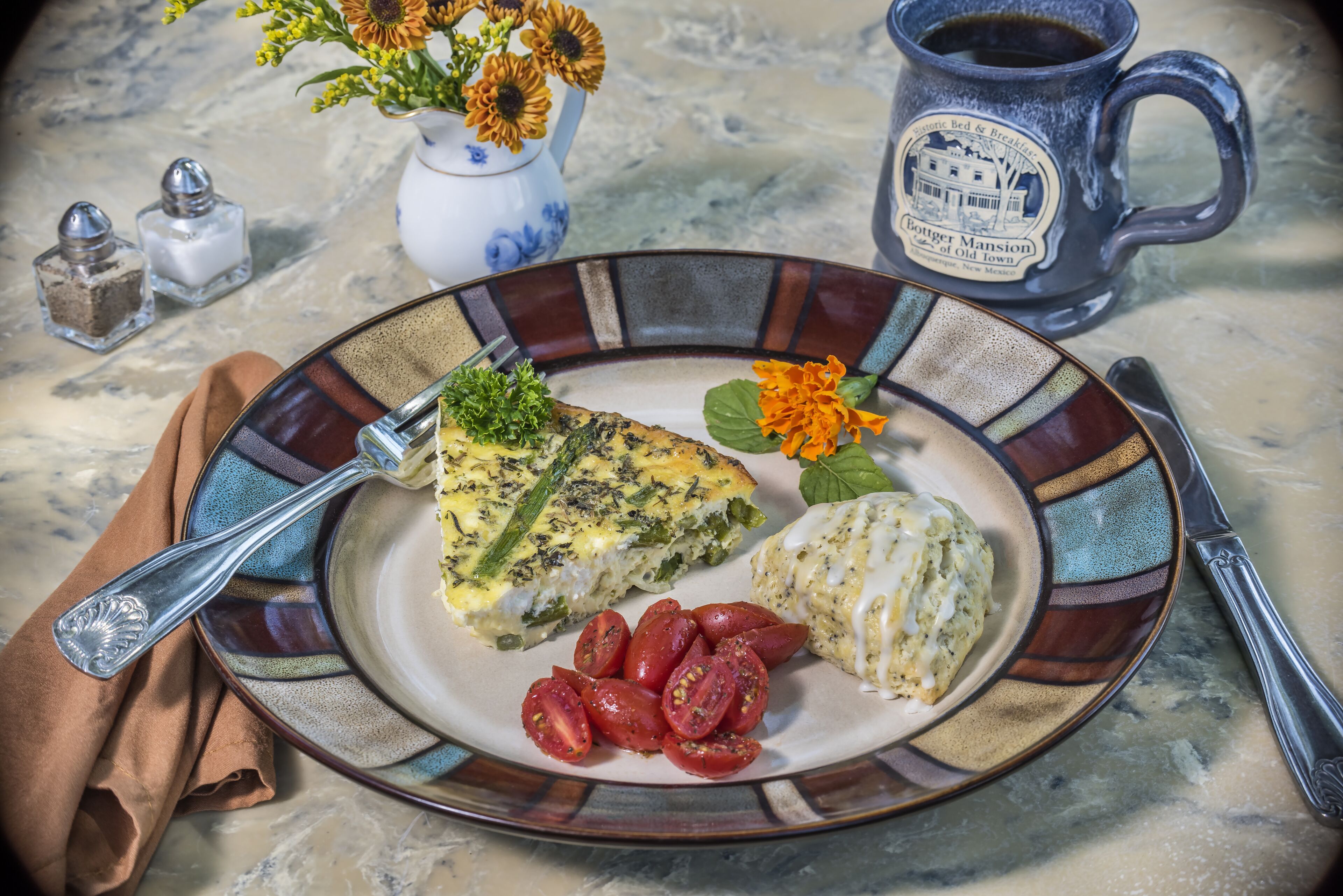 A plate with a quiche, a baked potato, and cherry tomatoes, accompanied by a mug and salt and pepper shakers on a marble table.