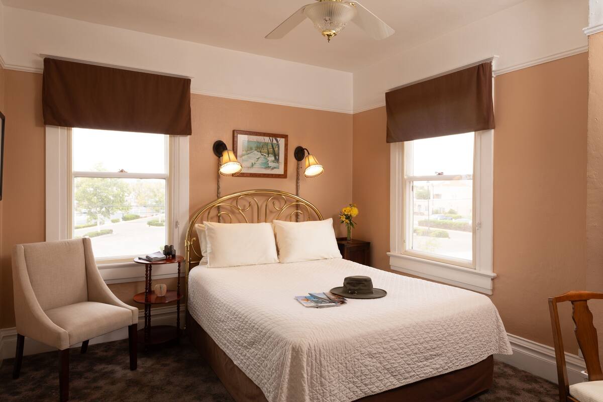 Une chambre à coucher bien rangée, avec un lit, deux lampes de chevet, une chaise, une table de chevet et une fenêtre donnant sur un beau paysage.