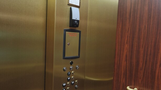 Interior de elevador com painel de controle, botão de emergência e porta de madeira.