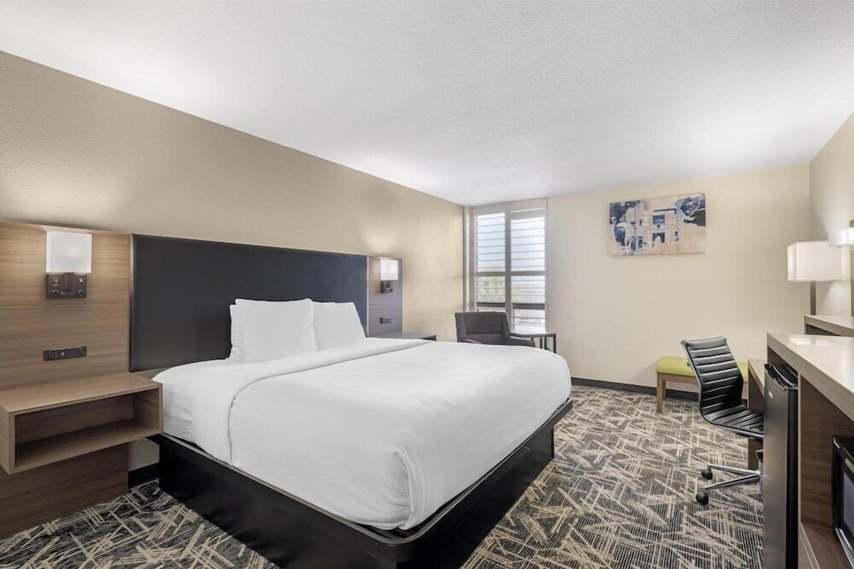 A hotel room with a bed, bedside table, lamp, chair, desk, and a painting on the wall.