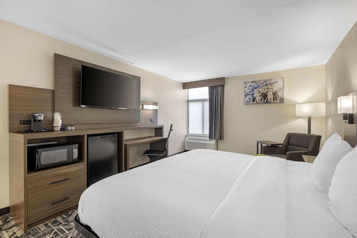 A hotel room with a large bed, a flat-screen TV, a desk with a coffee maker, a microwave, a chair, and a painting on the wall.