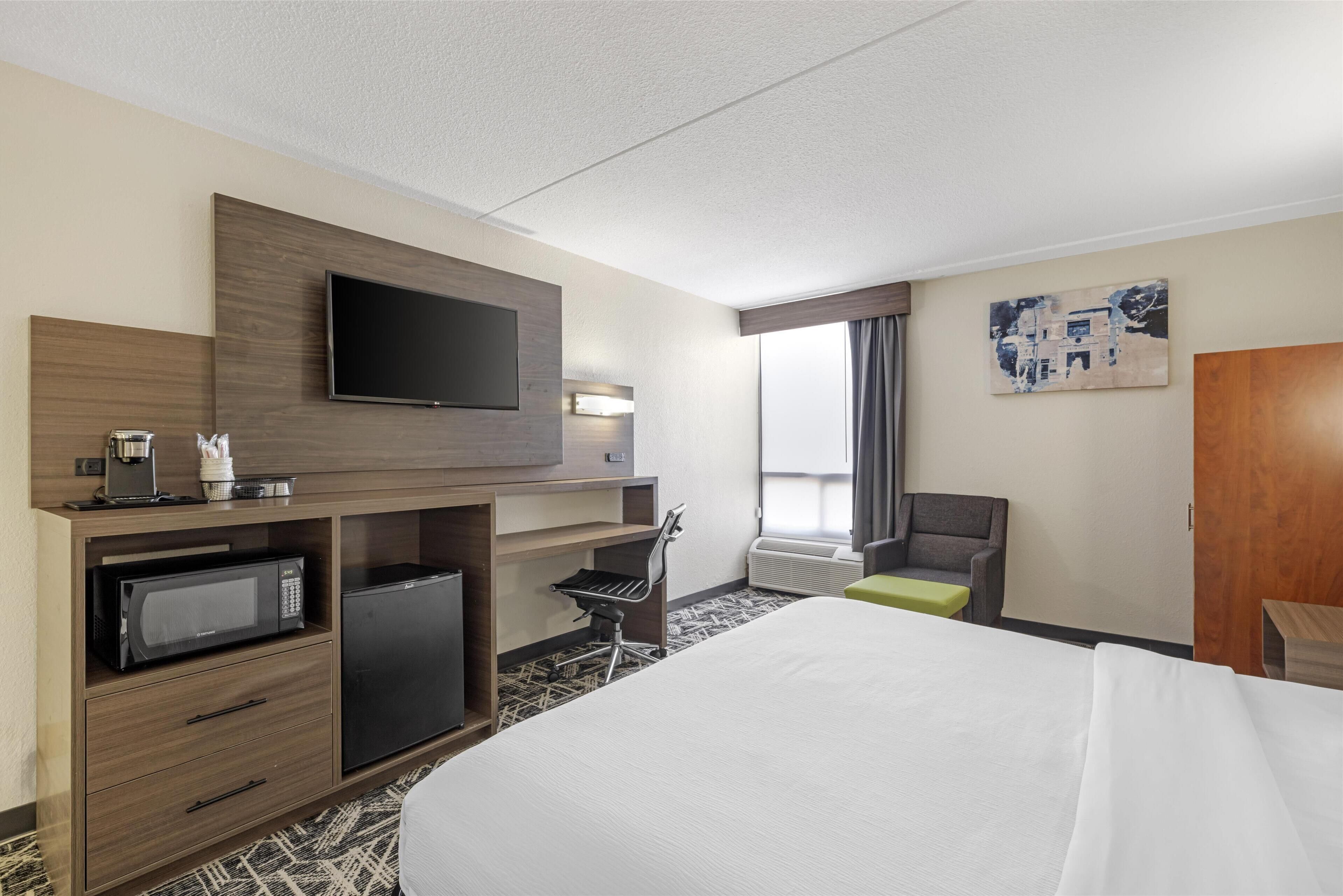 A hotel room with a bed, a desk with a flat-screen TV, a microwave, a mini-fridge, a chair, and a window with curtains.