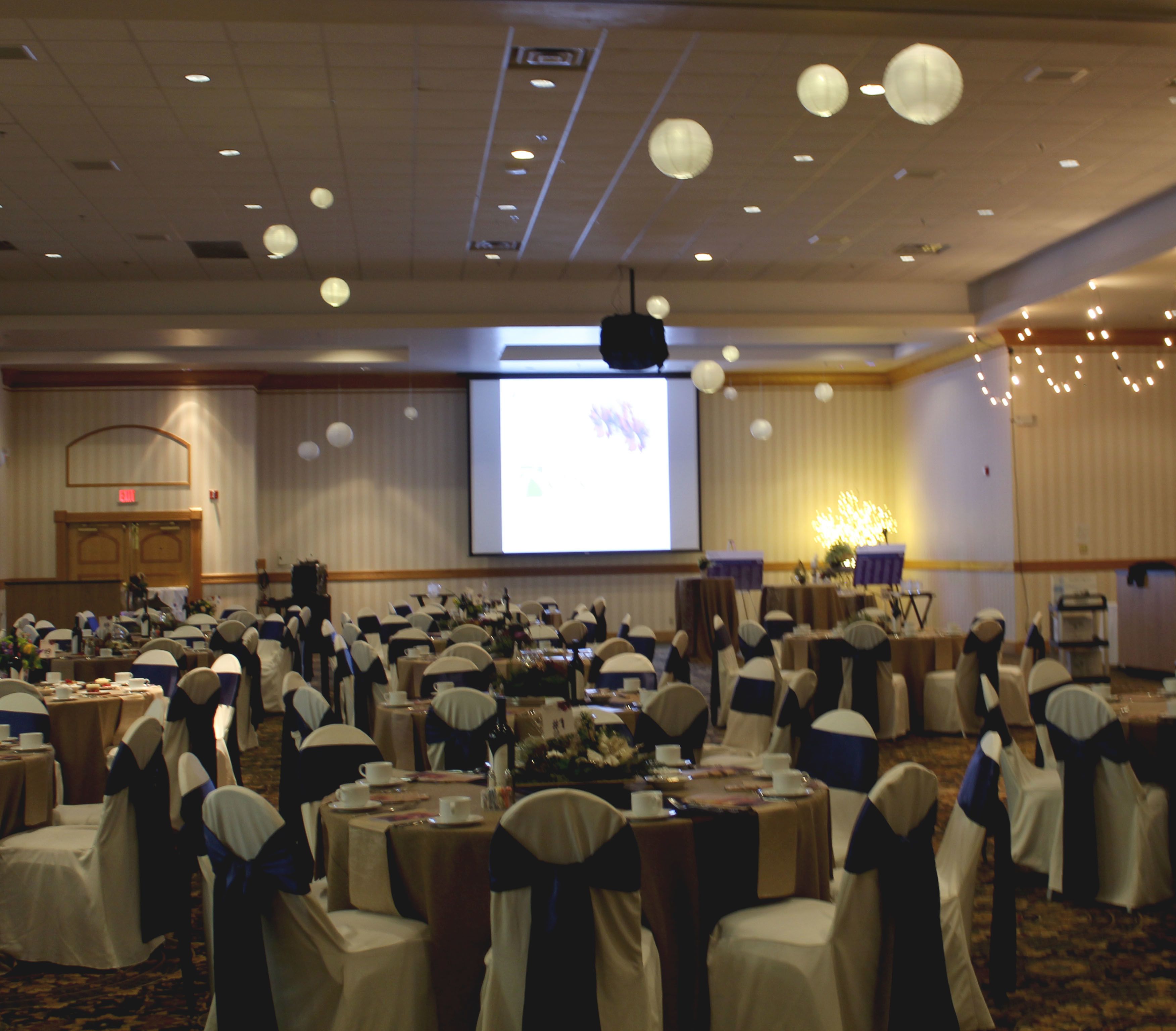 A banquet hall with round tables, chairs with sashes, a projector screen, and a stage with a podium.