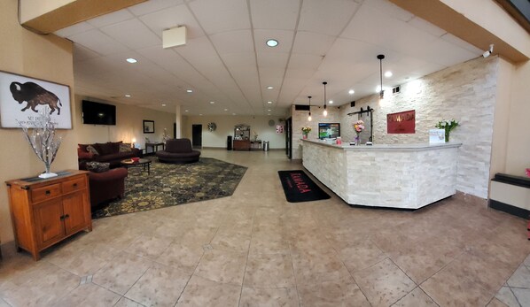 A hotel lobby with a reception desk, a reception area, a seating area with sofas, a decorative tree, and a carpet with a 'Ramada' sign.