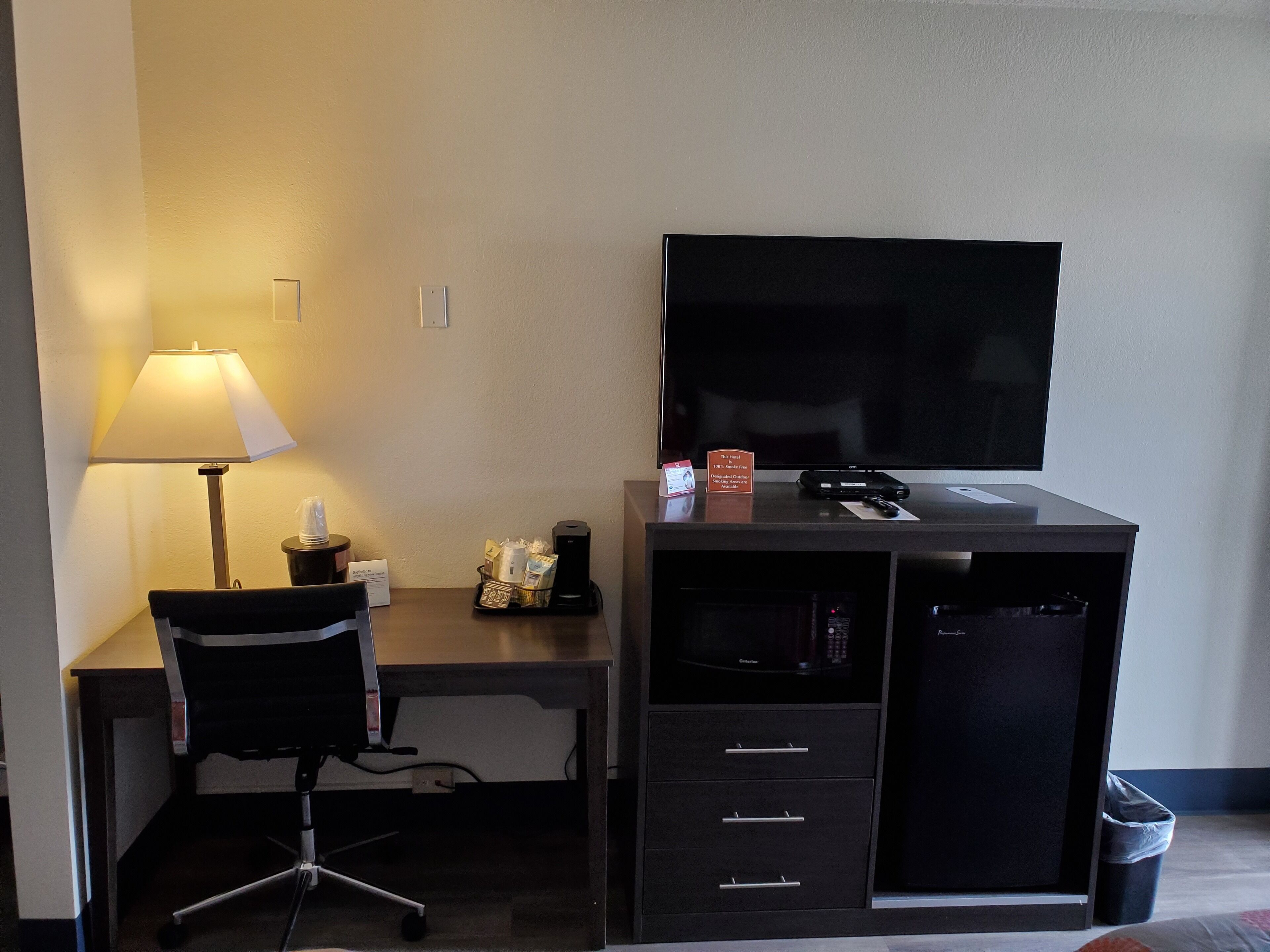 A hotel room with a desk, chair, TV, lamp, and a trash bin.