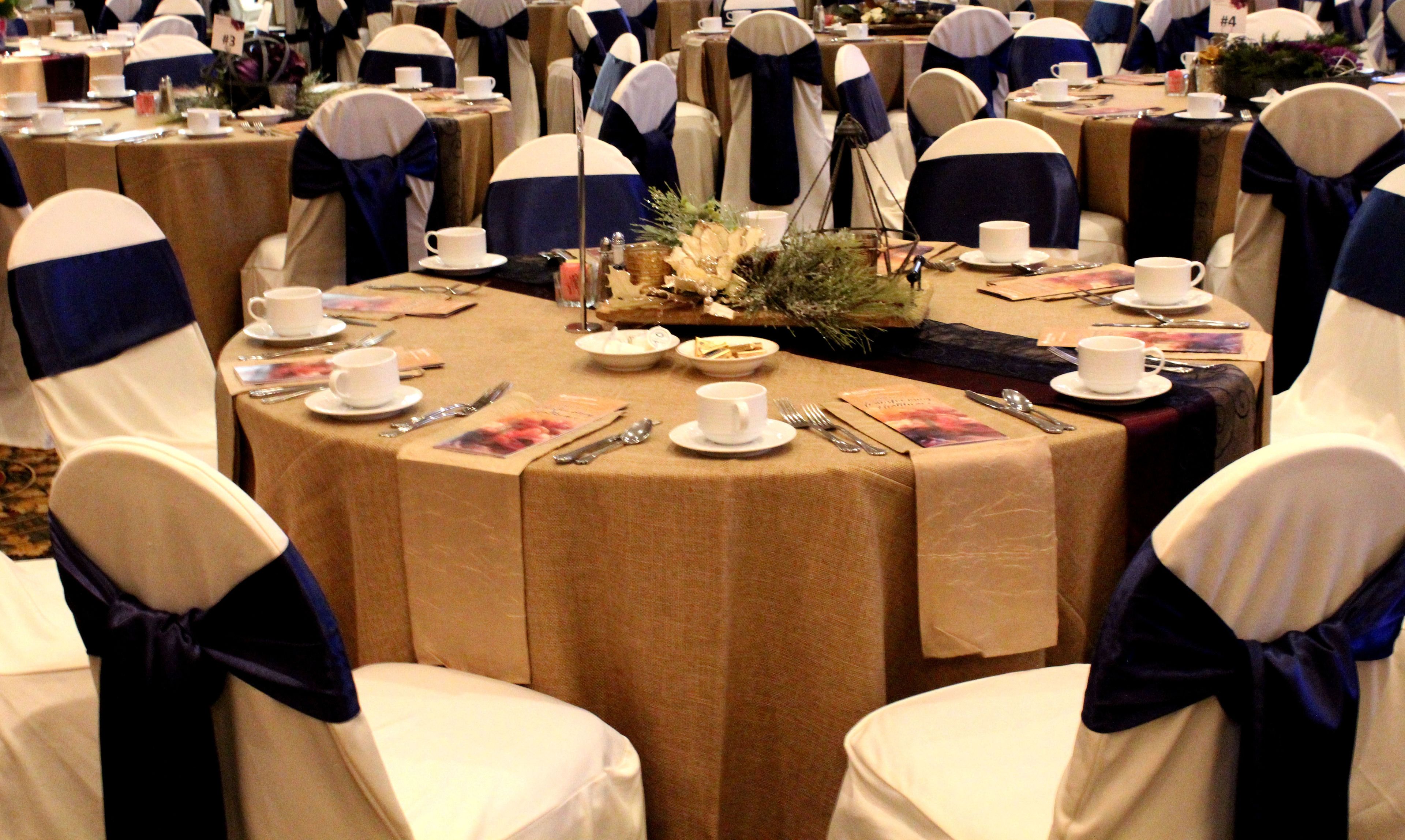 A round table set for a formal event with white chairs and blue sashes, surrounded by other similarly set tables.