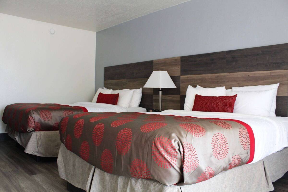 A hotel room with two beds, each with a red and grey patterned bedspread, white pillows, and a wooden headboard.