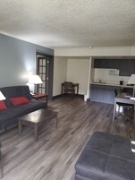 Una sala de estar con un sofá, una mesa de centro y un comedor con una silla y una mesa. Se ve una cocina con armarios y un microondas al fondo.