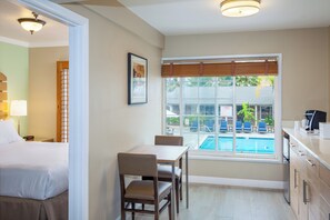 Una habitación de hotel con cama, una mesa de comedor con sillas, un ventanal con vista a una piscina y sillones de descanso, y una kitchenette.