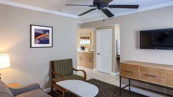 A hotel room with a ceiling fan, a television, a framed picture, a sofa, a chair, a small table, a cabinet, and a door leading to a bathroom.