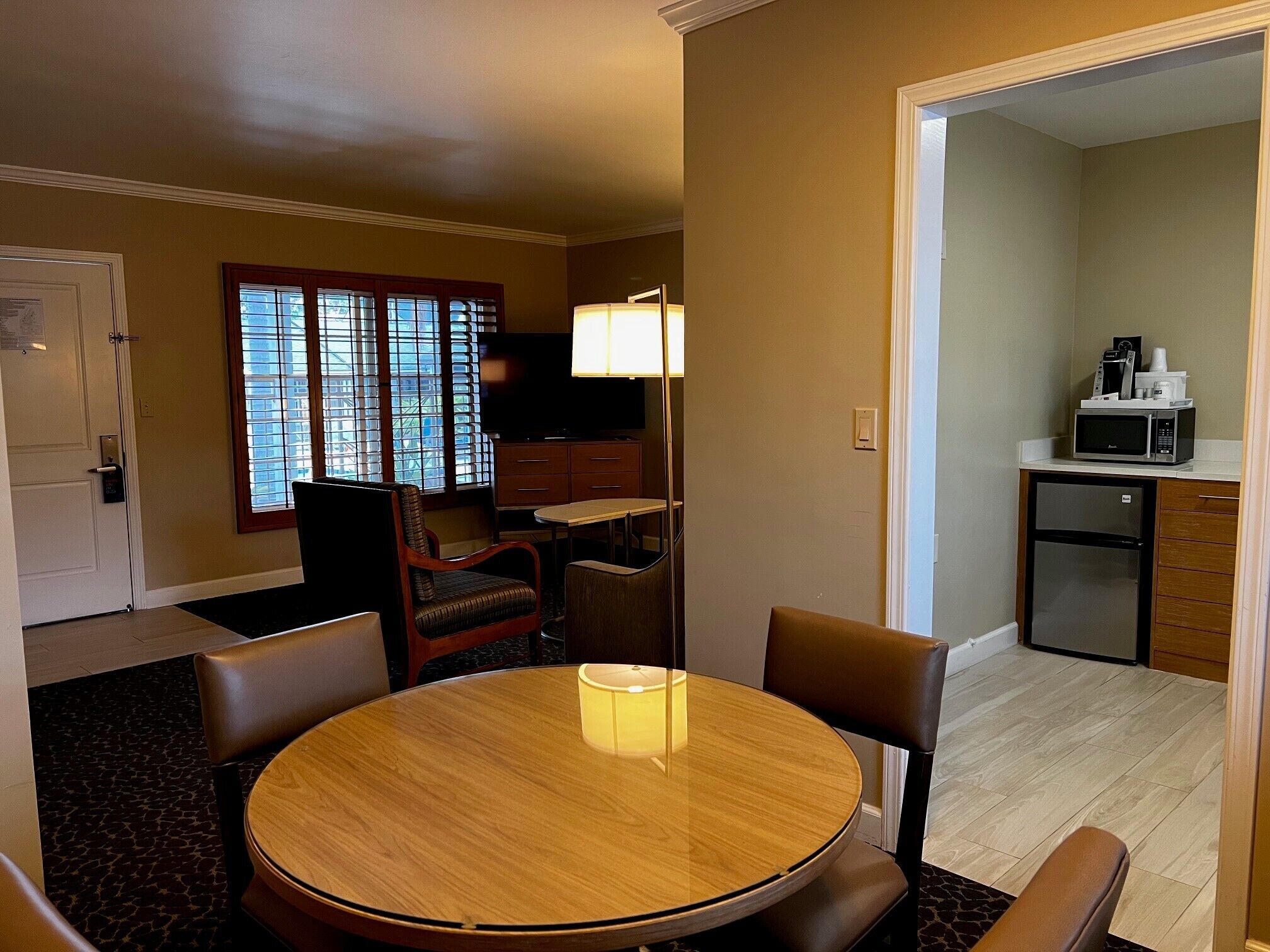 A hotel room with a round dining table, two chairs, a television, a lamp, a small kitchenette with a microwave, and a window with blinds.
