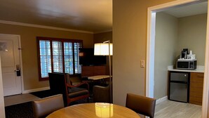 Habitación de hotel con una mesa de comedor redonda, dos sillas, televisor, lámpara, una pequeña kitchenette con microondas y ventana con persianas.