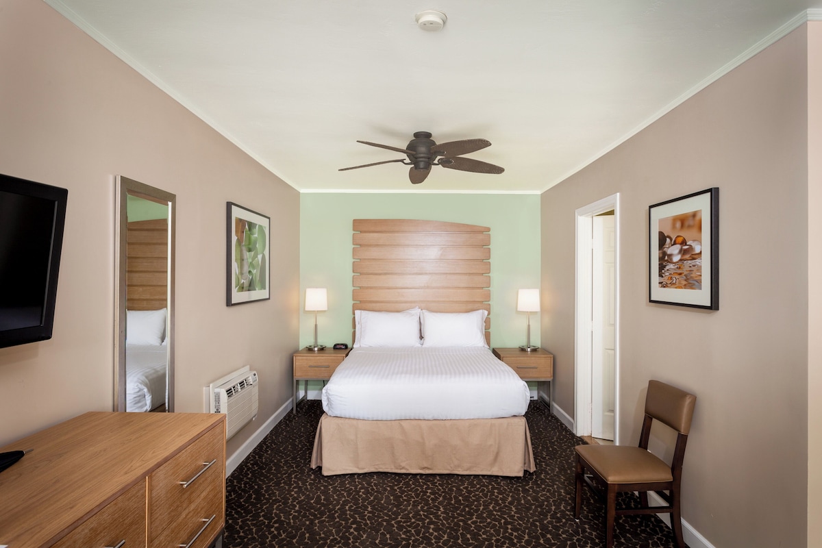 Une chambre d’hôtel équipée d’un lit, d’une télévision, d’un bureau, d’une chaise, d’un ventilateur de plafond et de tableaux encadrés accrochés aux murs.