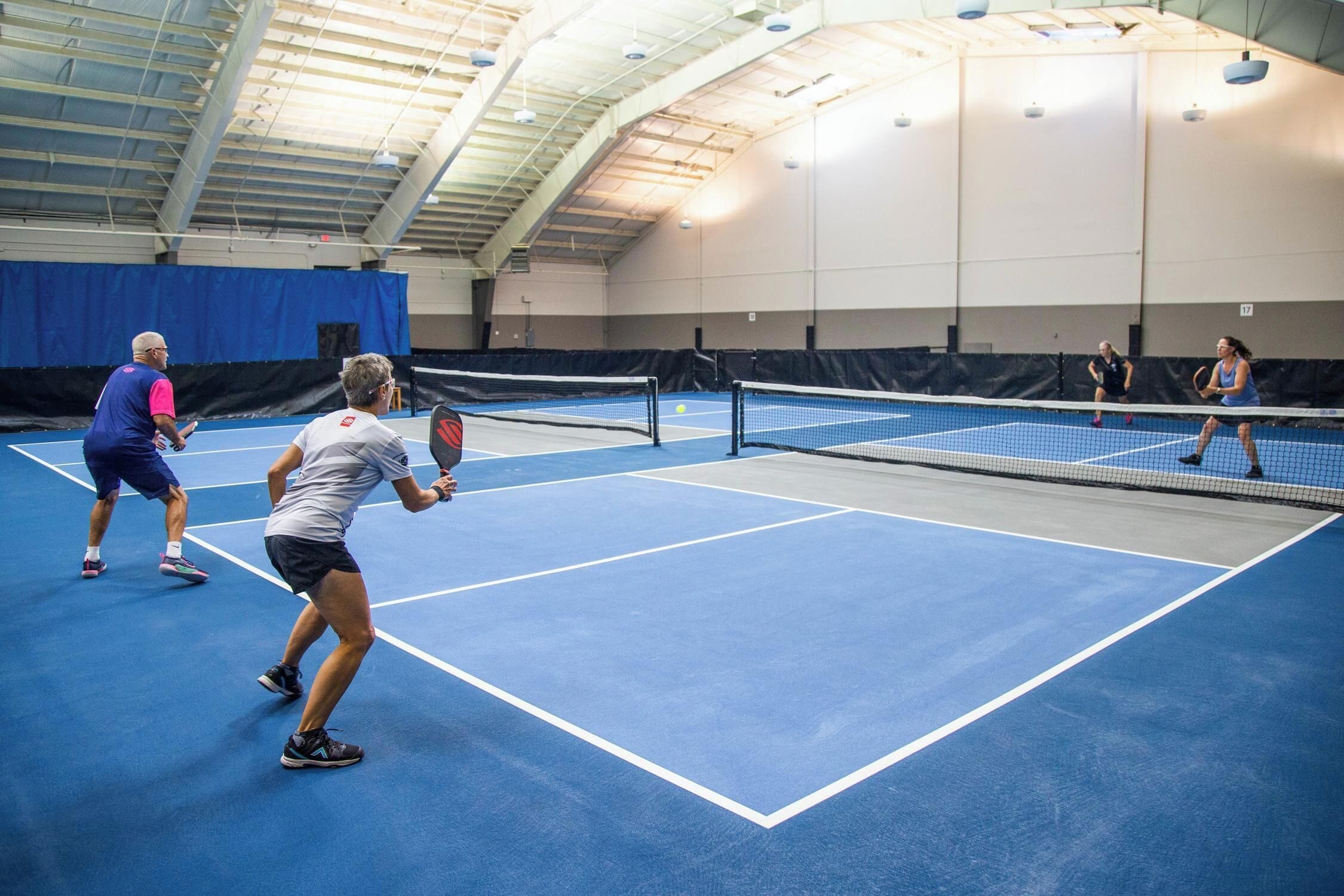 An indoor tennis court with a net, blue flooring, and a high ceiling with visible beams.