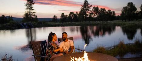 Una pareja sentada junto a una fogata al anochecer, con un lago al fondo.