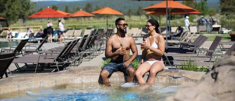 Una pareja sentada junto a una piscina con sillones y sombrillas al fondo.