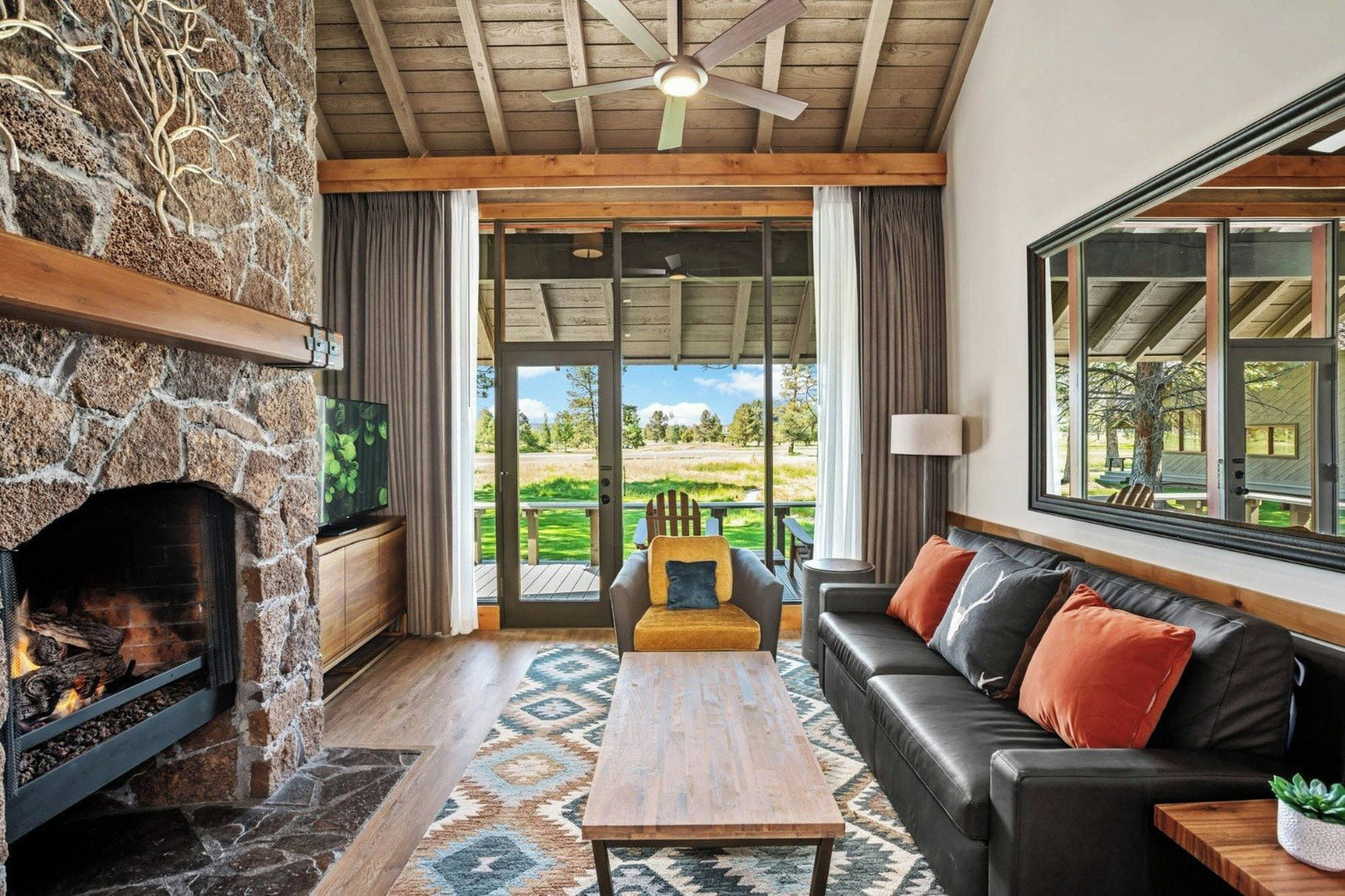A cozy living room with a stone fireplace, a sofa, a coffee table, and a view of a green landscape through large windows.