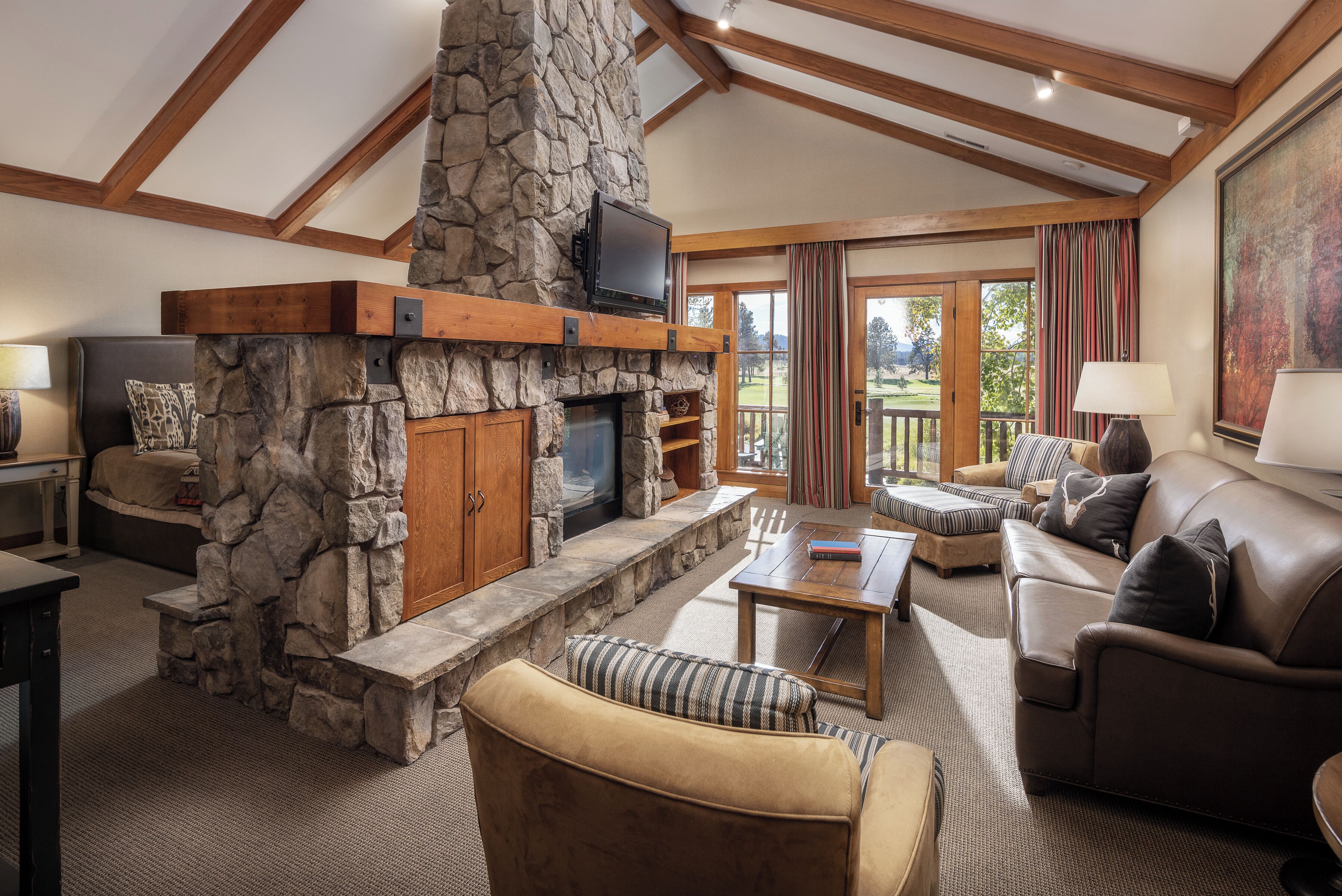 A stone fireplace, a sofa, a coffee table, and a television in a living room with wooden beams and a view to the outside.