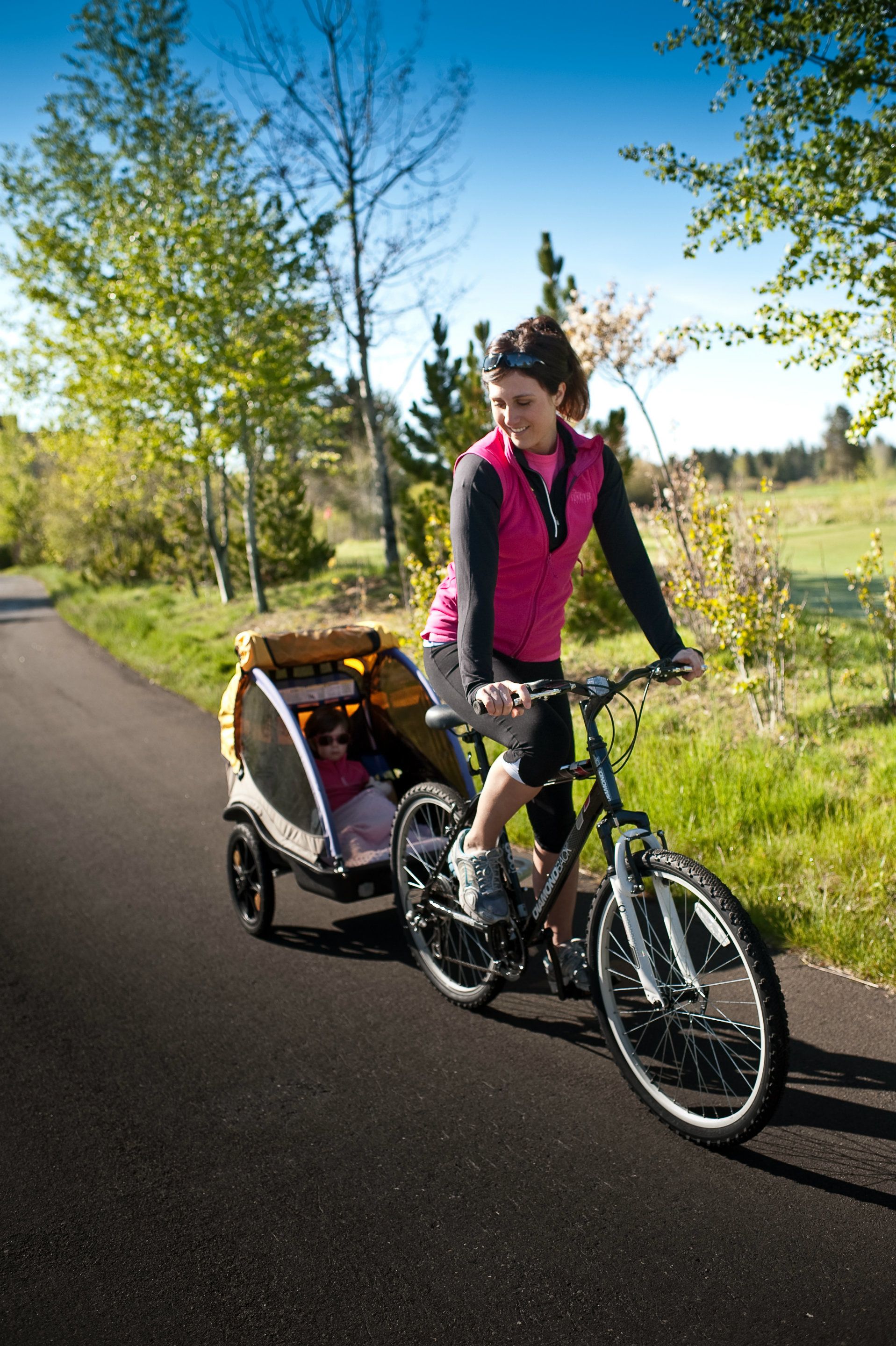 Une femme fait du vélo sur une piste cyclable, avec un enfant dans une remorque attachée au vélo.