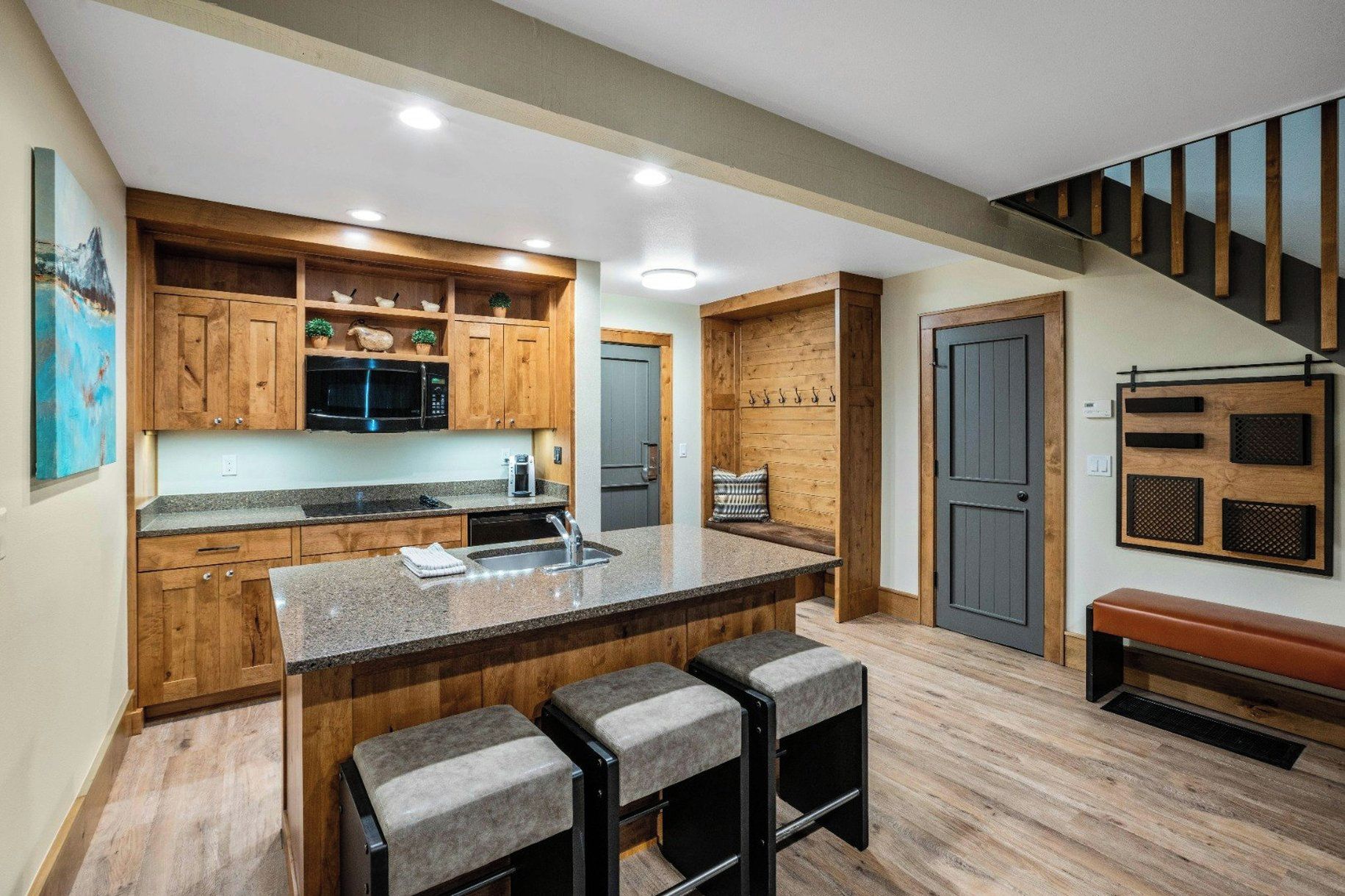 A modern kitchen with wooden cabinets, a granite countertop, and a built-in microwave. There are two bar stools and a bench.